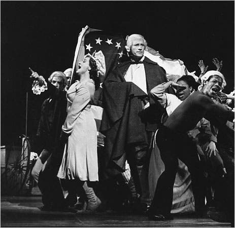 Ken Howard and Patricia Routledge stand alongside several dancers, mid-song. They are backed by an early American flag.